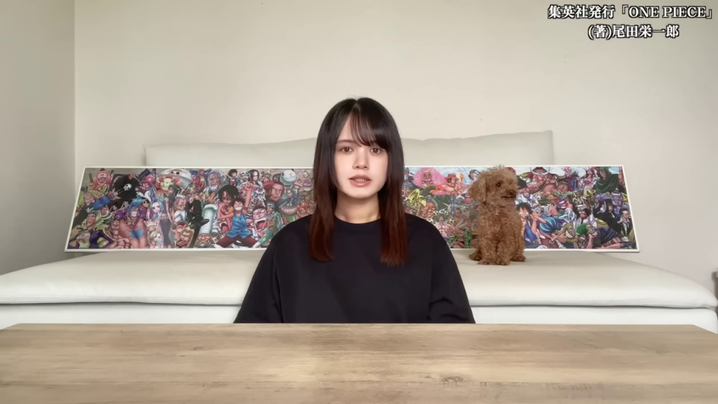 - 女性が座り、マンガ「ONE PIECE」の大きなイラストが飾られたソファの前で、カメラに向かって話している様子。犬もソファのそばにいる。この画像は、物語の舞台設定を示し、ONE PIECEの世界観への導入をスムーズにするために最適です。
