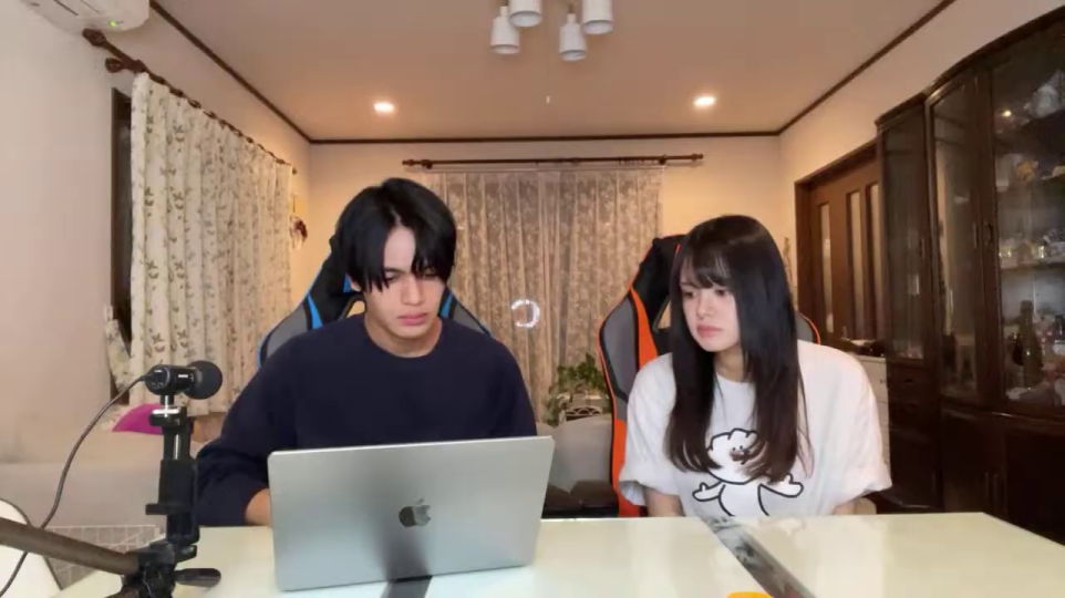 女性が男性の画面に集中する: 男性がMacBook Proの画面を真剣に見つめ、女性もスマートフォンを置いて体ごと画面の方を向き、集中している様子。このシーンは、二人が共通の関心事に没頭していることを示し、視聴者にもその情報への興味を促す。